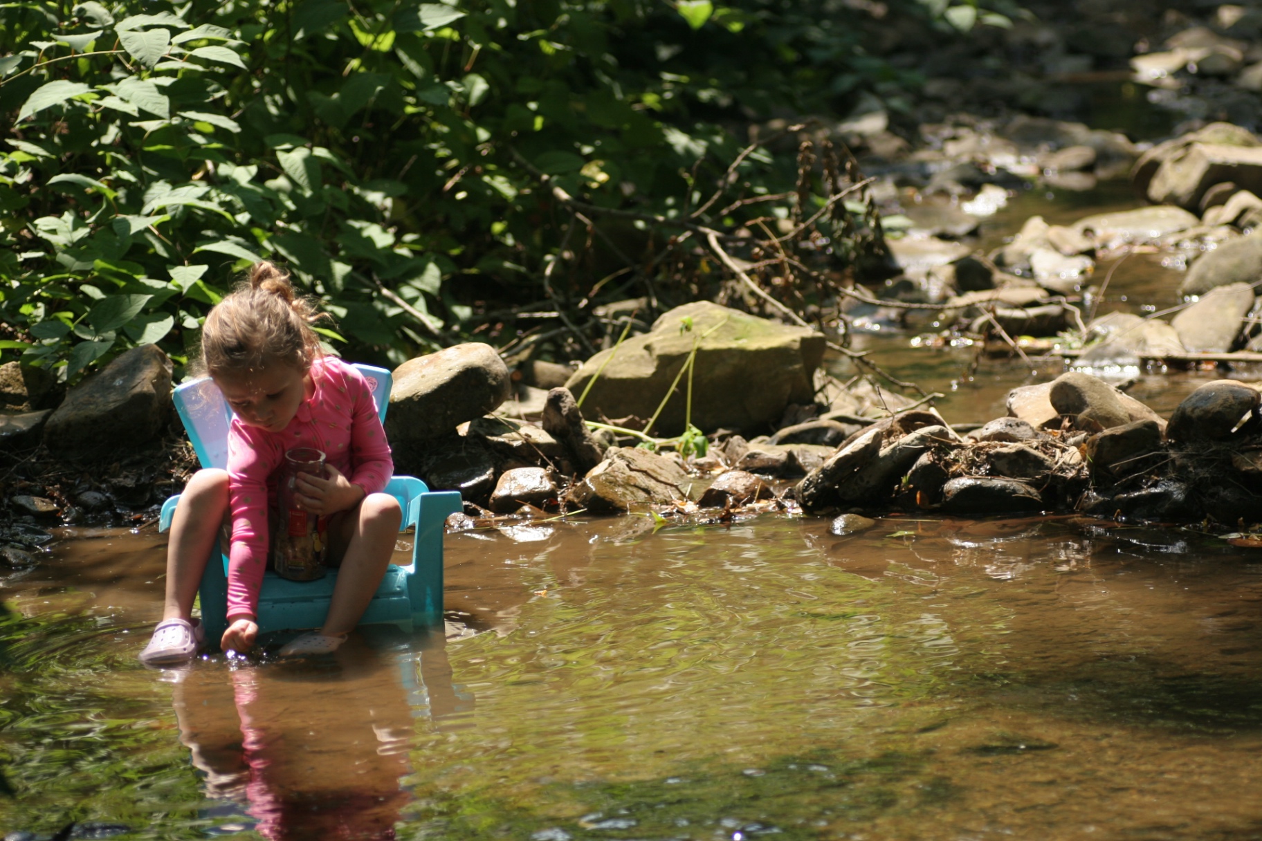 A child sitting in a small plastic chair in a shallow creek, holding a jar