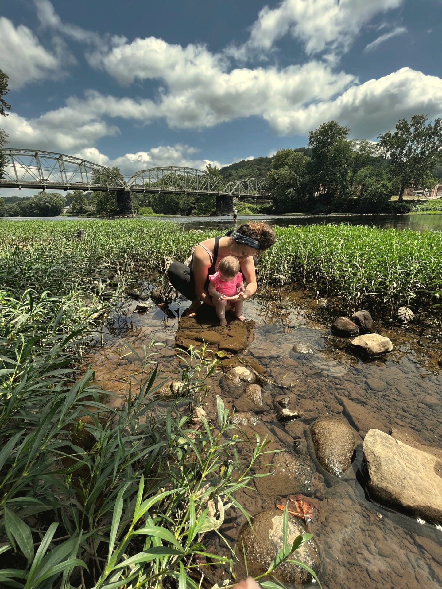 A mother crouched at the river's edge with her baby, a steel bridge and summer sky behind them